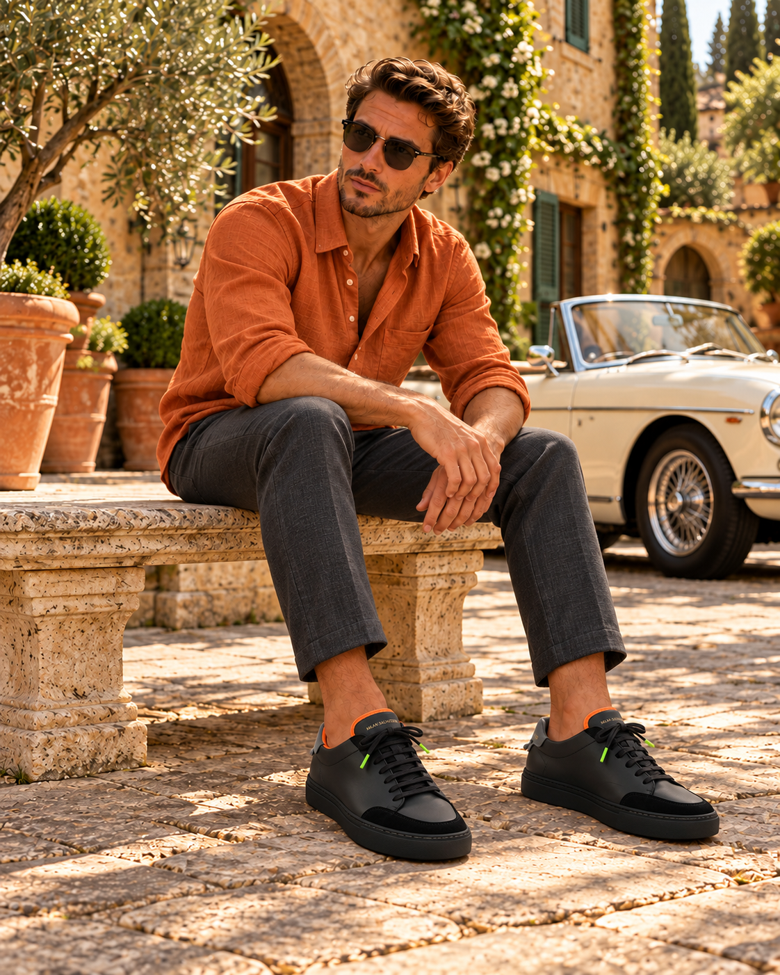 A man in a rust linen shirt and charcoal trousers seated on a stone bench beside a vintage convertible, wearing the Nero Everyday