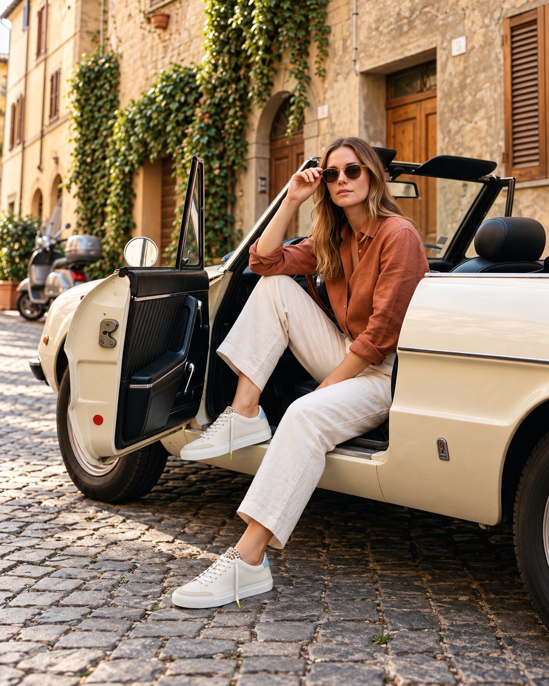 A woman in a terracotta linen shirt and cream trousers seated in a vintage convertible on a cobbled street, wearing the Cremino Everyday