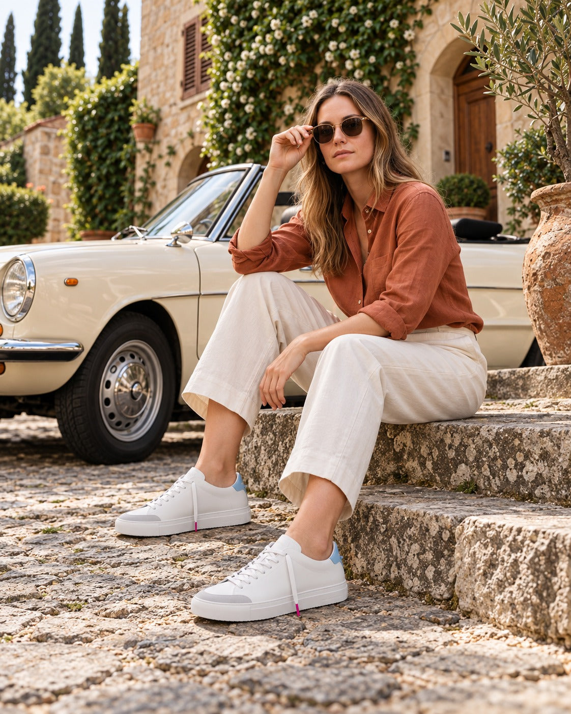 A woman in a terracotta linen shirt and cream trousers seated on stone steps beside a vintage convertible, wearing the Celeste Everyday